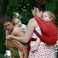 Egy édesanyának segít hordozókendőt kötni egy babáját szintén hordozó édesanya